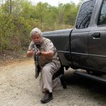 Bob Campbell crouching behind a pickup truck to provide concealment and cover