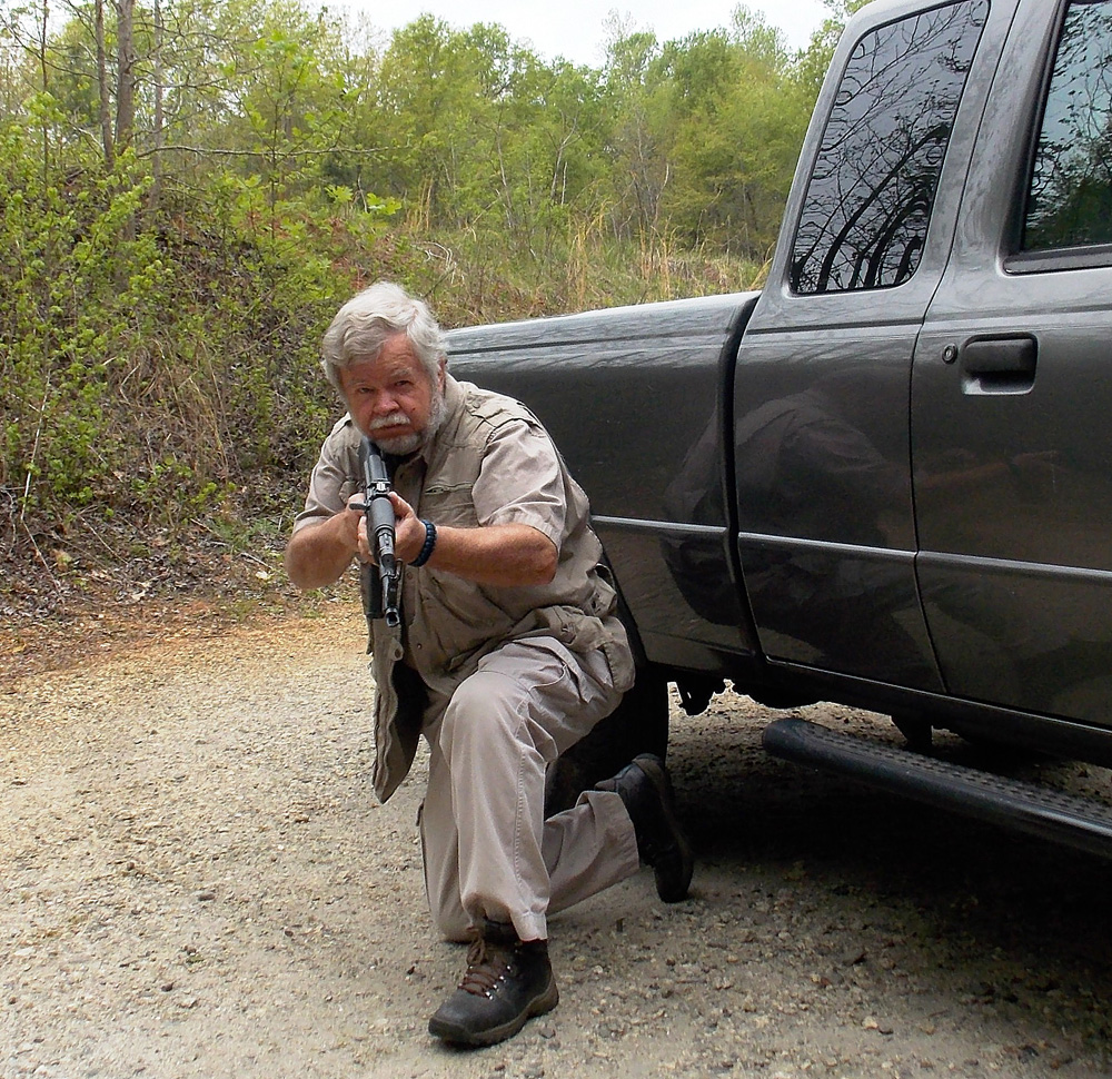 Firing a rifle from a kneeling position next to a truck for rifle marksmanship