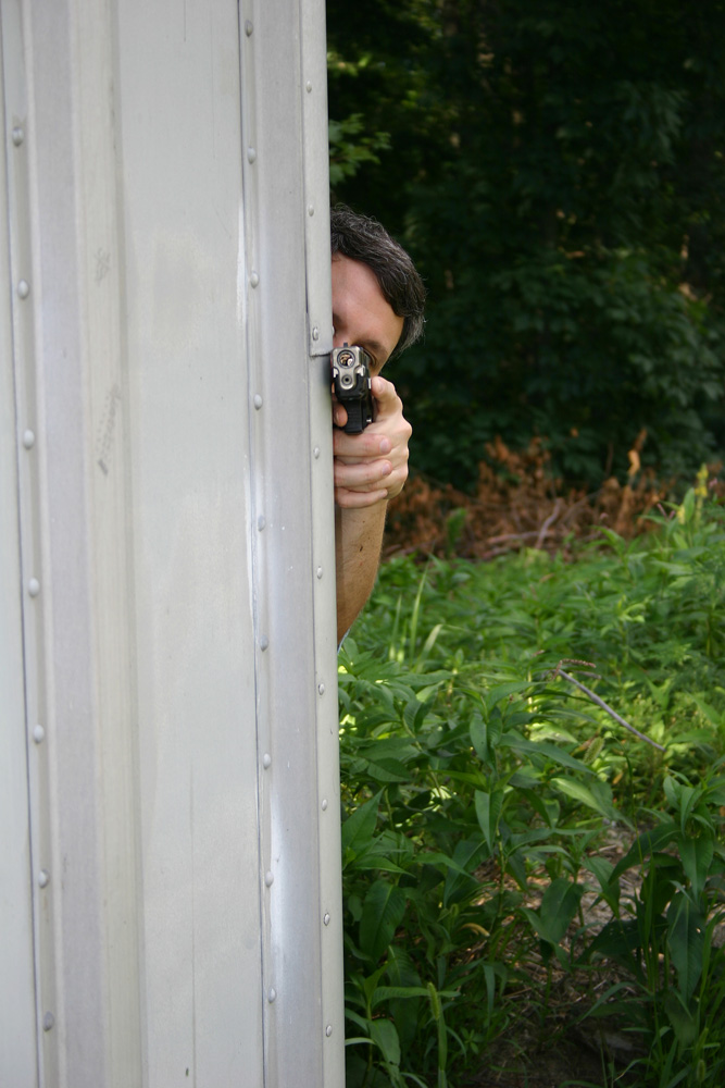 man shooting a pistol while using the corner of a metal shed as cover