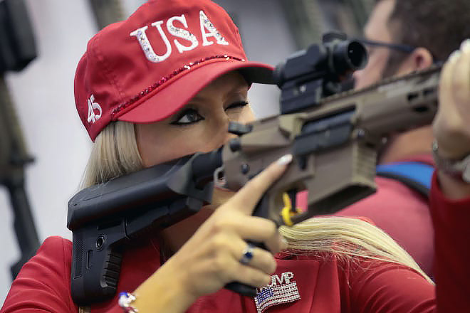 Woman holding an AR-15 wearing a ball cap with USA embroidered and a Trump 2020 logo on her shirt.