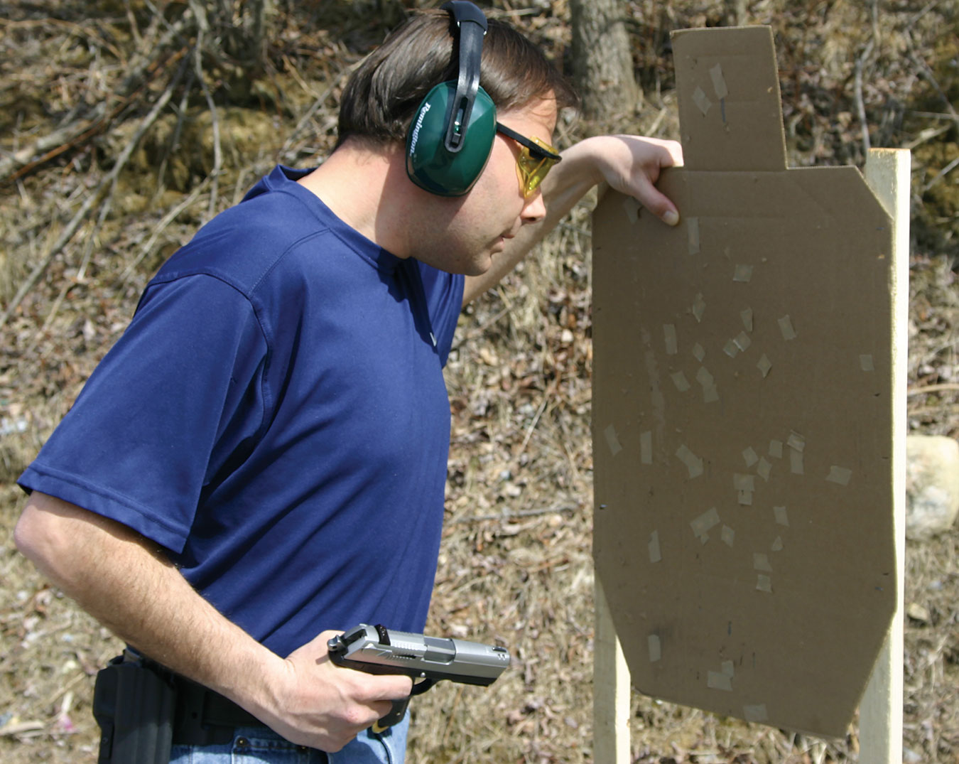 Man with one hand on a cardboard target and the other ready to fire his pistol from the retention position.